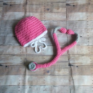 May include: A pink crocheted hat with a white trim and a matching pink and gray crocheted stethoscope. The stethoscope is shaped like a heart.