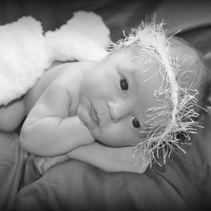 May include: A newborn baby wearing a white fluffy headband and a white fluffy nappy. The baby is lying on a white blanket and has its eyes closed.