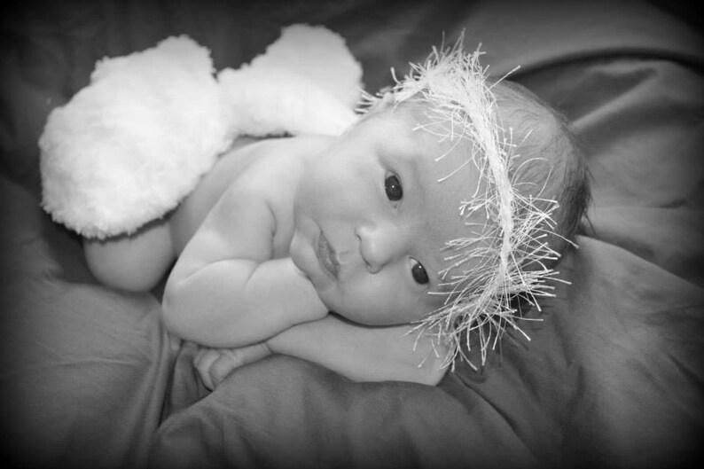 May include: A newborn baby wearing a white fluffy headband and a white fluffy nappy. The baby is lying on a white blanket and has its eyes closed.