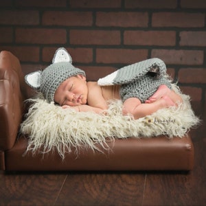 May include: A newborn baby wearing a gray crocheted wolf costume with a white tail and ears. The baby is sleeping on a white fluffy blanket on a brown leather couch.