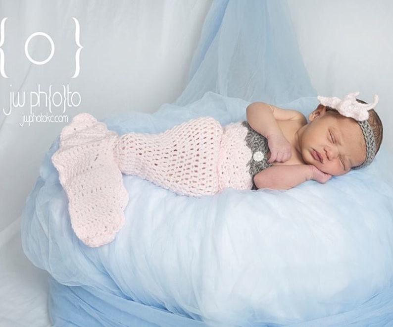May include: A newborn baby wearing a pink and gray crocheted mermaid costume, sleeping on a light blue fabric background.