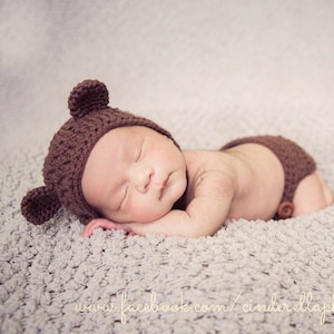 May include: A newborn baby wearing a brown crocheted bear hat and matching diaper cover, sleeping on a light gray fuzzy blanket.