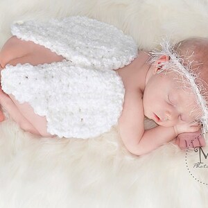 May include: A sleeping newborn baby wearing a white crocheted angel wing costume and a white crocheted headband. The baby is lying on a white fluffy surface.