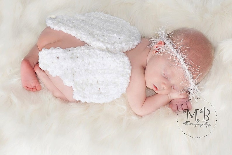 May include: A sleeping newborn baby wearing a white crocheted angel wing costume and a white crocheted headband. The baby is lying on a white fluffy surface.