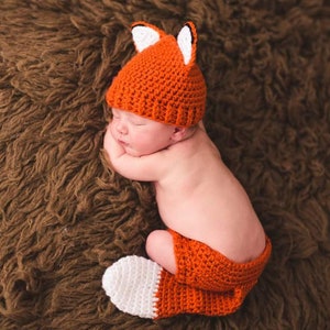 May include: A newborn baby wearing a crocheted fox costume. The costume is orange with white accents and includes a hat with ears and a tail.