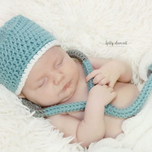 May include: A baby wearing a light blue crocheted hat with a white trim and a light blue crocheted stethoscope. The baby is sleeping on a white fluffy blanket.