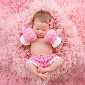 May include: A newborn baby wearing a pink crocheted boxing outfit, including a headband, gloves, and shorts, is sleeping on a pink fluffy blanket.