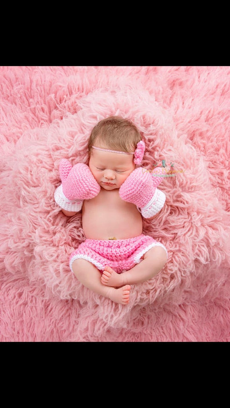 May include: A newborn baby wearing a pink crocheted boxing outfit, including a headband, gloves, and shorts, is sleeping on a pink fluffy blanket.