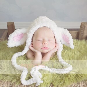 May include: A newborn baby wearing a white crocheted lamb hat with pink ears, lying on a green fuzzy blanket in a woven basket.