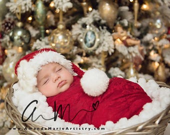 May include: A sleeping baby wearing a red and white Santa hat and a red blanket with white trim, lying in a wicker basket decorated with white pom-poms. The basket is in front of a Christmas tree decorated with ornaments and lights. The text "www.AmandaMarieArtistry.com" is visible at the bottom of the image.