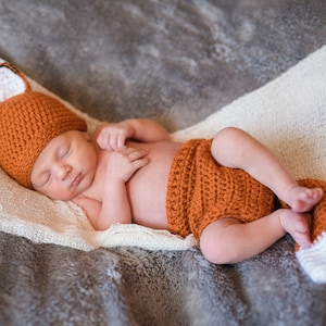 May include: A newborn baby wearing a crocheted fox costume, sleeping on a white and gray blanket. The costume is orange and white with a fox tail.