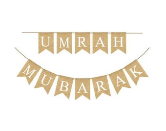 May include: A burlap banner with the words "Umrah Mubarak" in white letters. The banner is strung together with a white string.