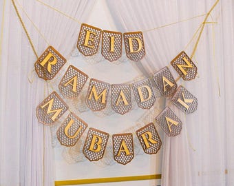 May include: A gold banner with the words "Eid Mubarak Ramadan" hanging on a white curtain.
