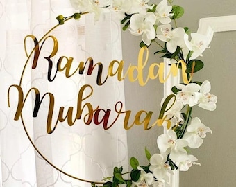 May include: A gold-coloured circular Ramadan Mubarak sign decorated with white orchids and green leaves. The sign hangs in front of a white curtain, creating a festive and elegant decoration.