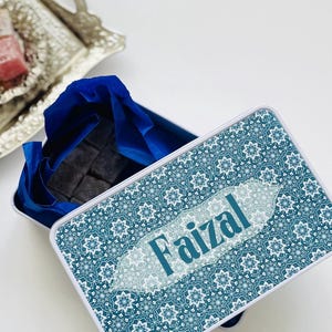 Peut inclure: Boîte cadeau rectangulaire ouverte, ornée d'un motif géométrique turquoise et blanc. Elle contient du papier de soie bleu et des friandises foncées. Le nom "Faizal" est imprimé sur le couvercle en turquoise clair.