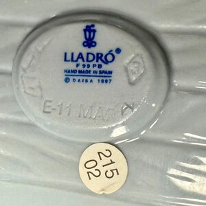 May include: A close-up of a white ceramic piece with the LLADR&Oacute; logo and text that reads "HAND MADE IN SPAIN" and "DAISA 1997". A small sticker with the numbers "215 02" is also visible.