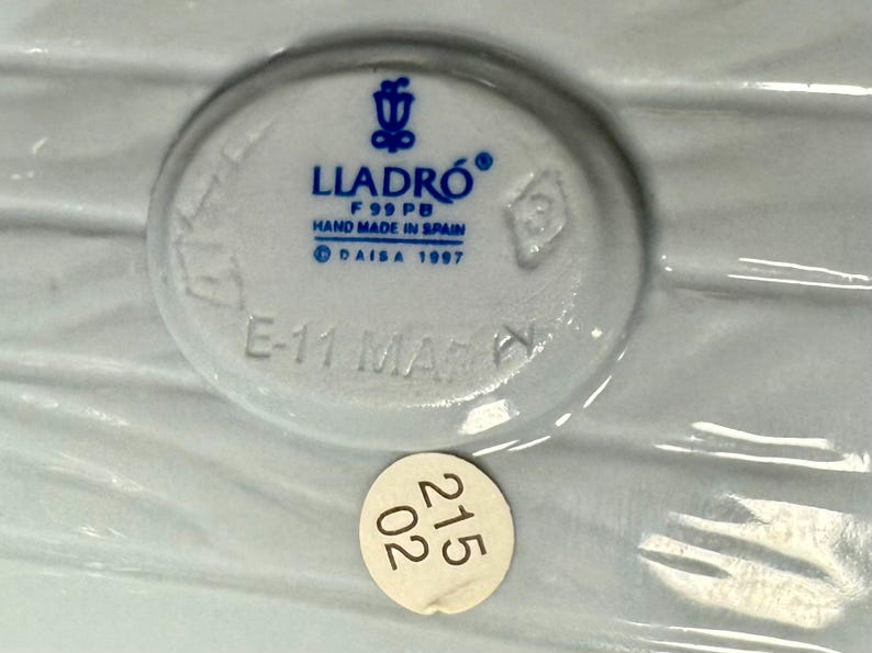 May include: A close-up of a white ceramic piece with the LLADR&Oacute; logo and text that reads "HAND MADE IN SPAIN" and "DAISA 1997". A small sticker with the numbers "215 02" is also visible.