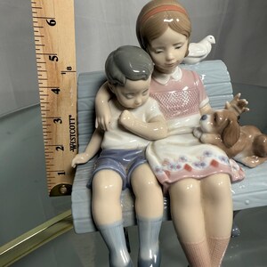 May include: A porcelain figurine depicting two children and a dog sitting on a bench. The girl wears a pink dress, and the boy wears blue shorts. A white dove sits on the bench behind them. The figurine is approximately 6 inches tall.