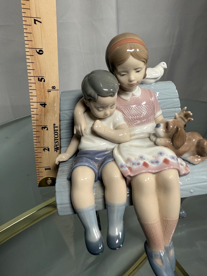 May include: A porcelain figurine depicting two children and a dog sitting on a bench. The girl wears a pink dress, and the boy wears blue shorts. A white dove sits on the bench behind them. The figurine is approximately 6 inches tall.
