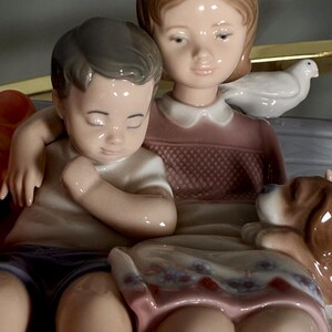 May include: A porcelain figurine depicting a girl and a boy seated together, with a small dog and a dove. The girl wears a pink dress, and the boy wears a white shirt and purple shorts. The figurine has a glossy finish.