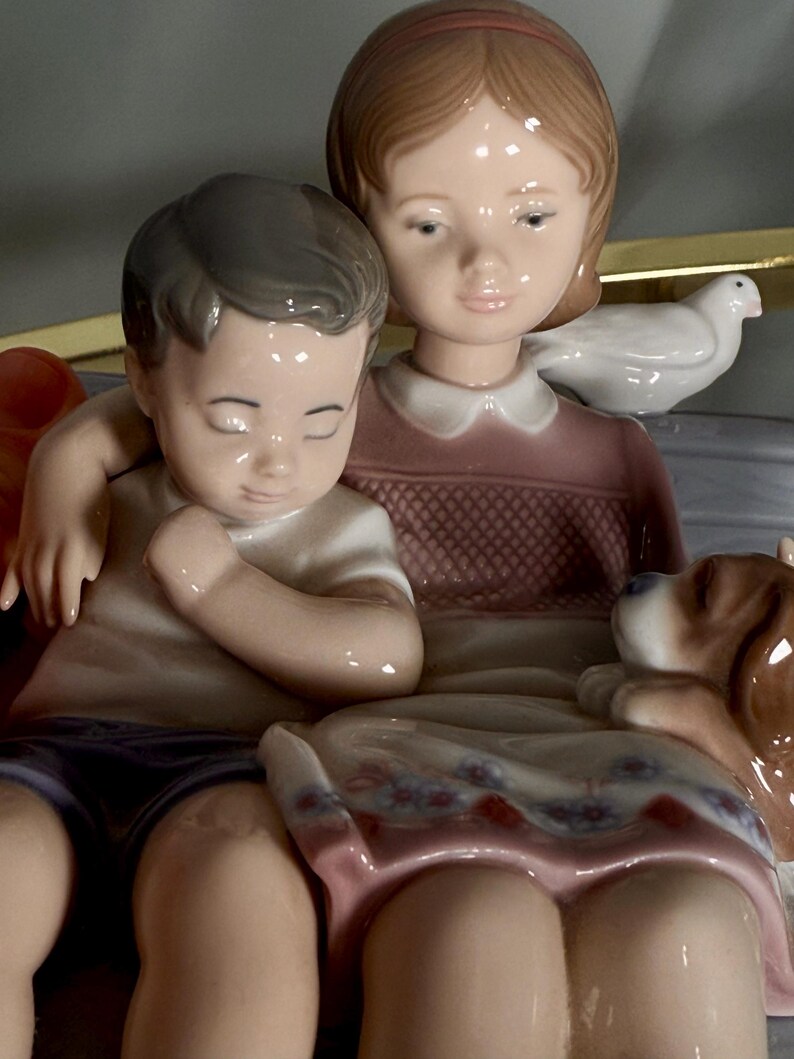May include: A porcelain figurine depicting a girl and a boy seated together, with a small dog and a dove. The girl wears a pink dress, and the boy wears a white shirt and purple shorts. The figurine has a glossy finish.