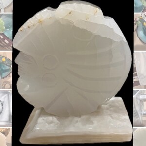 May include: White alabaster sculpture of a stylized face with intricate details. The sculpture is carved in a rounded shape and sits on a white base.