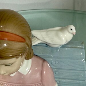 May include: A porcelain figurine featuring a young girl in a pink dress, with brown hair and an orange hairband. A white dove rests on a light blue bench. The figurine has a glossy finish and is set against a light green background.
