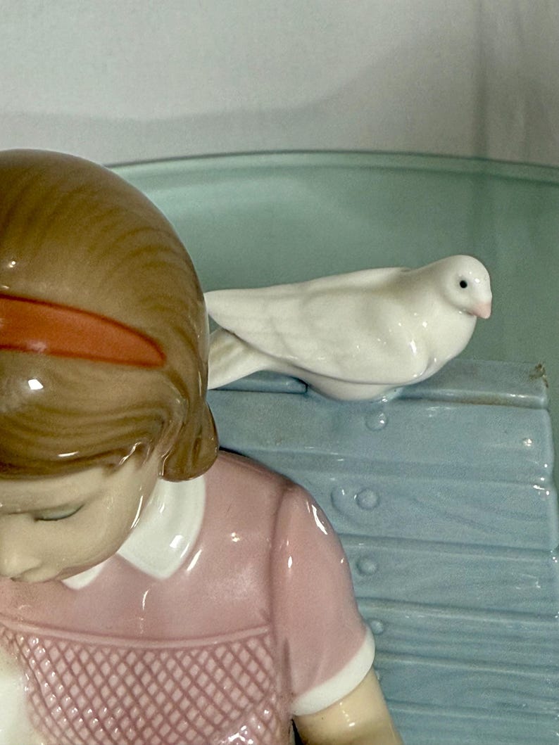 May include: A porcelain figurine featuring a young girl in a pink dress, with brown hair and an orange hairband. A white dove rests on a light blue bench. The figurine has a glossy finish and is set against a light green background.