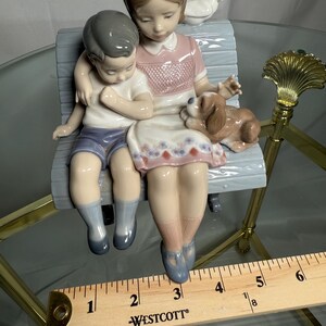 May include: A porcelain figurine depicting a girl and a boy seated on a bench, with a small dog and a dove. The girl wears a pink dress, and the boy is in blue shorts. The bench is light blue, and the figurine has a glossy finish.