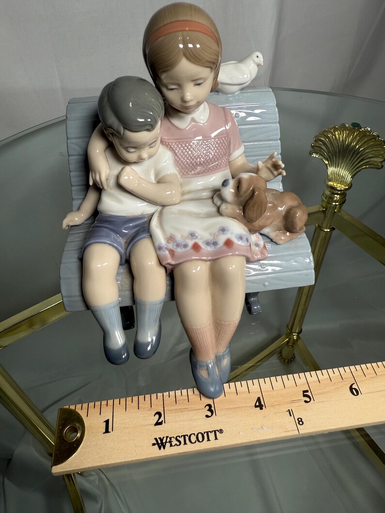 May include: A porcelain figurine depicting a girl and a boy seated on a bench, with a small dog and a dove. The girl wears a pink dress, and the boy is in blue shorts. The bench is light blue, and the figurine has a glossy finish.
