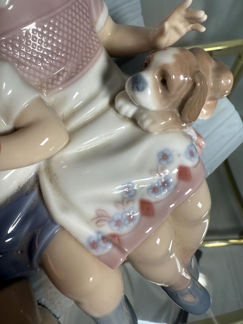May include: A porcelain figurine of a child with a puppy. The child is wearing a pink top and blue shorts. The puppy is brown and white. The figurine has a glossy finish and is highly detailed.