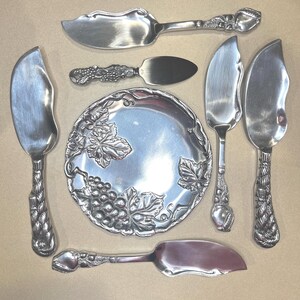 May include: A set of six silver-plated serving pieces with grapevine and grape designs. The set includes a small serving dish, a butter knife, and four cheese knives.
