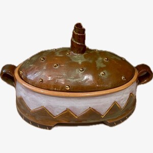 May include: A handcrafted ceramic casserole dish with a lid. The dish features a brown and green speckled glaze, gold accents, and a white and brown geometric design. It has two handles and a decorative spiral on the lid.