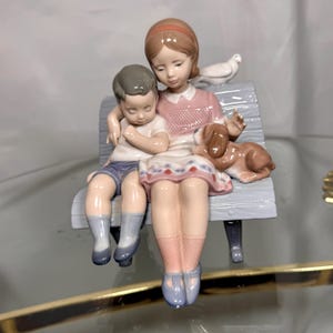 May include: A porcelain figurine depicting two children and a dog sitting on a bench. The girl wears a pink dress and the boy wears a white shirt and blue shorts. A brown dog rests beside them, and a white dove sits on the bench.