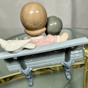 May include: A ceramic figurine depicting two figures seated on a light blue bench. One figure has light brown hair, the other has gray hair. A white dove sits on the bench. The figures are wearing pink and white clothing.