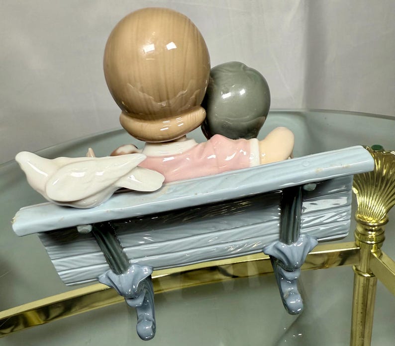 May include: A ceramic figurine depicting two figures seated on a light blue bench. One figure has light brown hair, the other has gray hair. A white dove sits on the bench. The figures are wearing pink and white clothing.