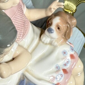 May include: A porcelain figurine depicting a child with a puppy. The child is wearing a pink and white dress with blue accents. The puppy is brown and white. The figurine has a glossy finish and is set against a light blue background.