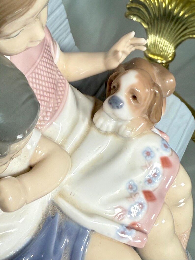 May include: A porcelain figurine depicting a child with a puppy. The child is wearing a pink and white dress with blue accents. The puppy is brown and white. The figurine has a glossy finish and is set against a light blue background.