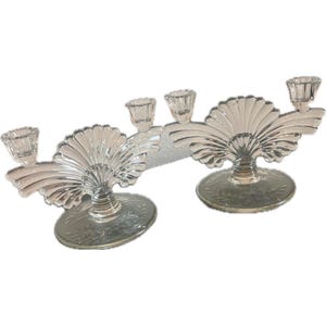 May include: A pair of clear glass candle holders with a fan-shaped design. Each holder has three candle cups and a circular base.