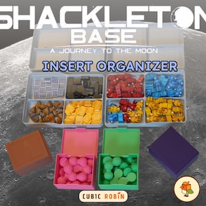 May include: A clear plastic insert organiser for the Shackleton Base board game, with compartments filled with game pieces in various colours. The text "INSERT ORGANIZER" is displayed. Additional coloured boxes are also shown.