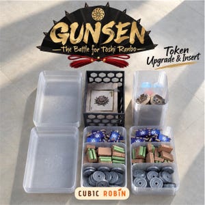 May include: A board game organiser set for Gunsen: The Battle for Toshi Ranbo, featuring clear plastic trays, a black hexagonal-patterned box, and various game tokens including blue banners, green and brown blocks, and grey circular coins.