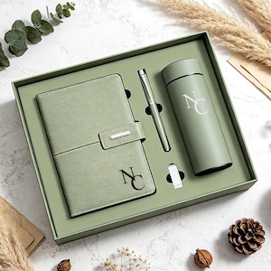 May include: A sage green gift set in a matching box containing a textured notebook with a silver clasp, a matching ballpoint pen, and a cylindrical insulated bottle. Both the notebook and the bottle feature the monogrammed letters NC in black.