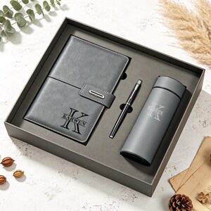 May include: A grey gift set containing a rectangular notebook with a magnetic closure, a black ballpoint pen, and a cylindrical insulated tumbler. The notebook and tumbler are both personalised with the letter K and the name KARWEN.