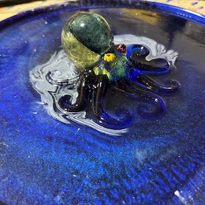 Pode incluir: Uma figura de polvo de vidro com tentáculos azuis e uma cabeça verde e amarela. O polvo está sentado em uma superfície azul e branca.