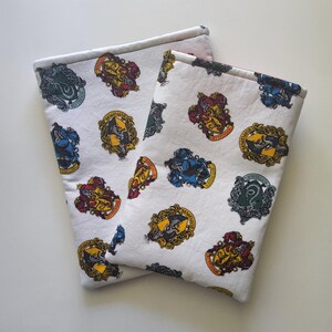 May include: Two white fabric book sleeves featuring a repeating pattern of colorful crests. The crests are in gold, red, blue, green, and yellow. The sleeves are folded over and appear to be made of cotton.