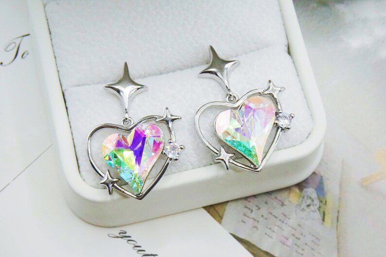 Pu&ograve; includere: Un paio di orecchini a forma di cuore color argento con centri di cristallo iridescenti multicolore. Ogni orecchino presenta un accento a forma di stella nella parte superiore e piccoli dettagli a stella attorno al cuore. Un piccolo cristallo trasparente &egrave; incastonato su ogni orecchino.