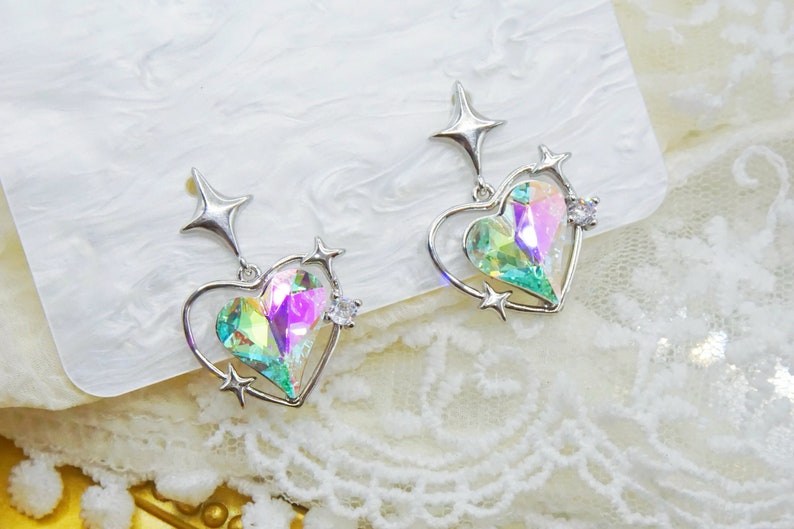 Pu&ograve; includere: Un paio di orecchini a forma di cuore color argento con centri di cristallo iridescenti multicolore. Ogni orecchino presenta una parte superiore a forma di stella e piccoli accenti a stella, con un abbellimento di cristallo trasparente.