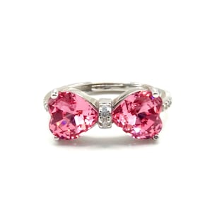 May include: A silver ring with a bow design, featuring two heart-shaped, pink gemstones. The bow's centre has a small, clear gemstone. The ring is set against a white background.
