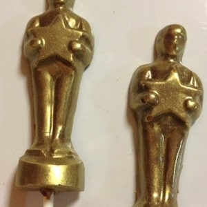 May include: Two gold-colored, statuette-shaped lollipops. Each lollipop features a figure holding a star, resembling an award trophy. The lollipops are on white sticks, perfect for a themed party or event.