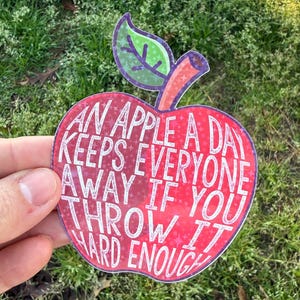 May include: A red apple-shaped sticker with the text "AN APPLE A DAY KEEPS EVERYONE AWAY IF YOU THROW IT HARD ENOUGH." The sticker has a green leaf and a pink stem. The sticker is held in front of a grassy background.